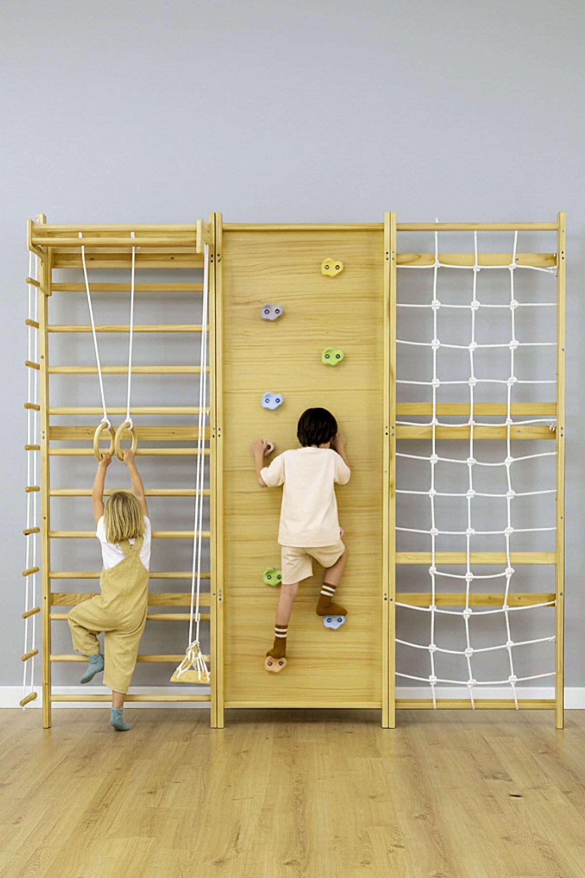 Two children play on a wooden modular jungle gym featuring climbing holds, a rope ladder, and gymnastic rings. The warm-toned wood and neutral walls create a bright, inviting play space.