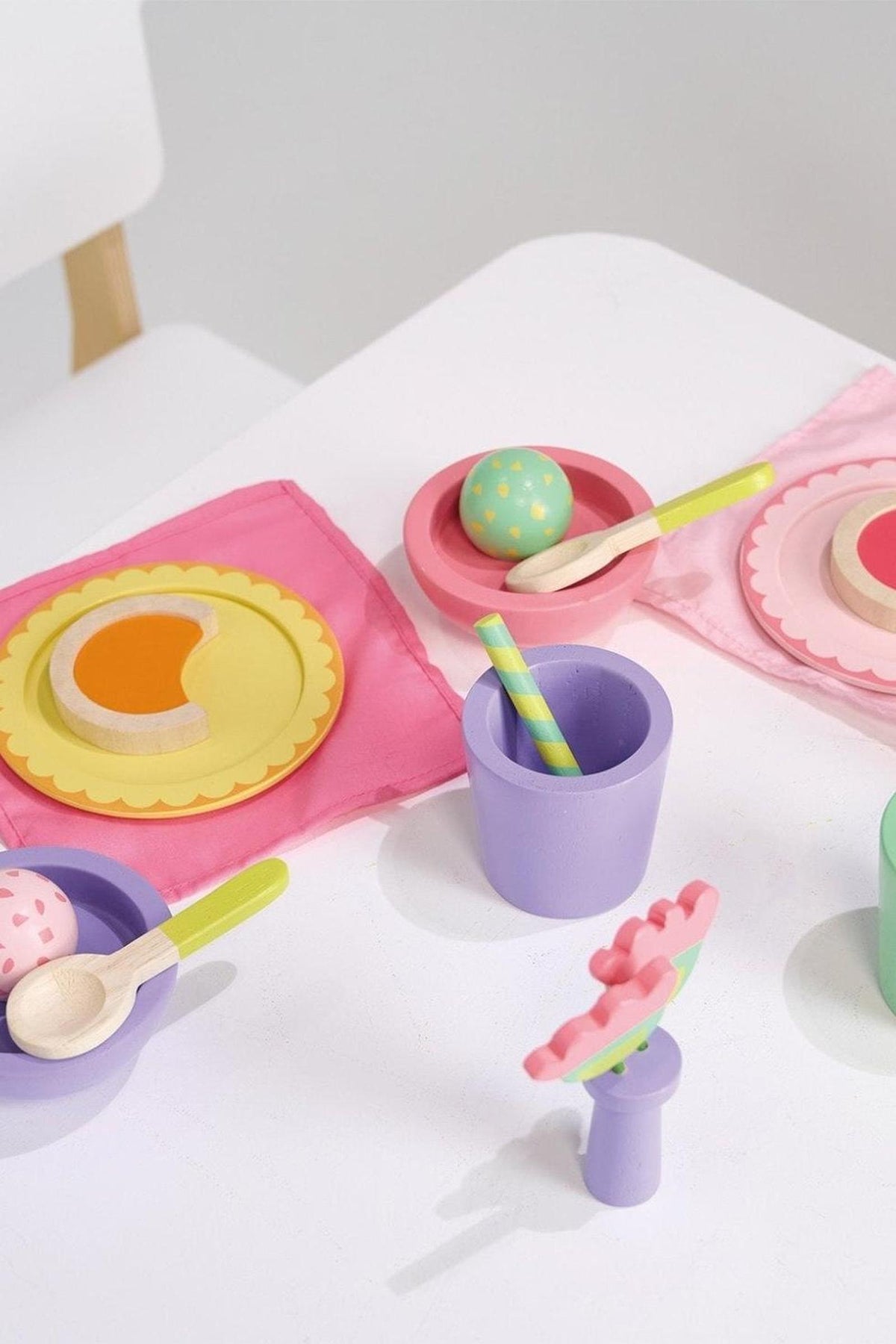 Colorful wooden toy table setting with plates, bowls, cups, and cutlery on a white surface with pink napkins.