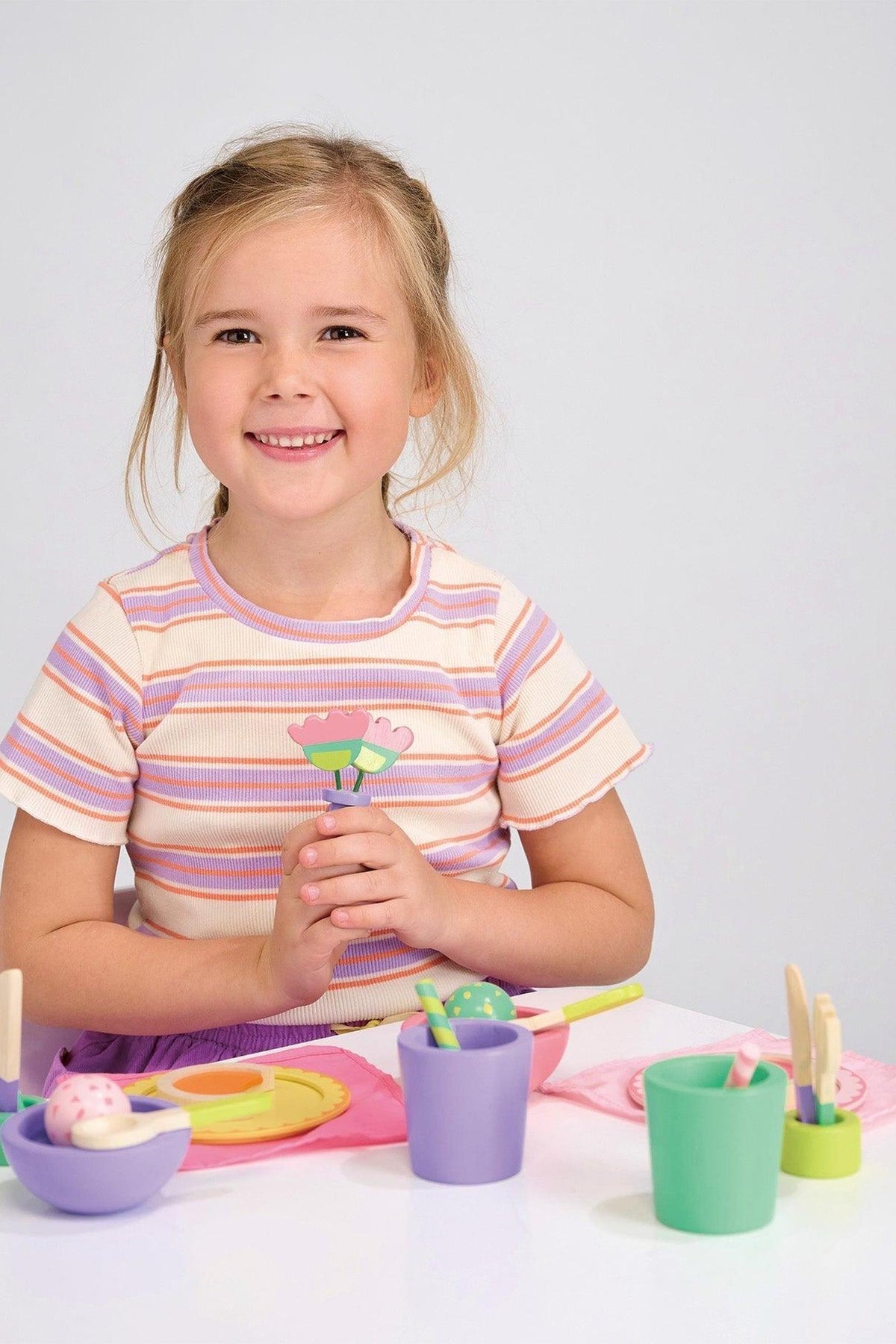 A smiling child is sitting at a table with colorful wooden toy cups and plates in front.
