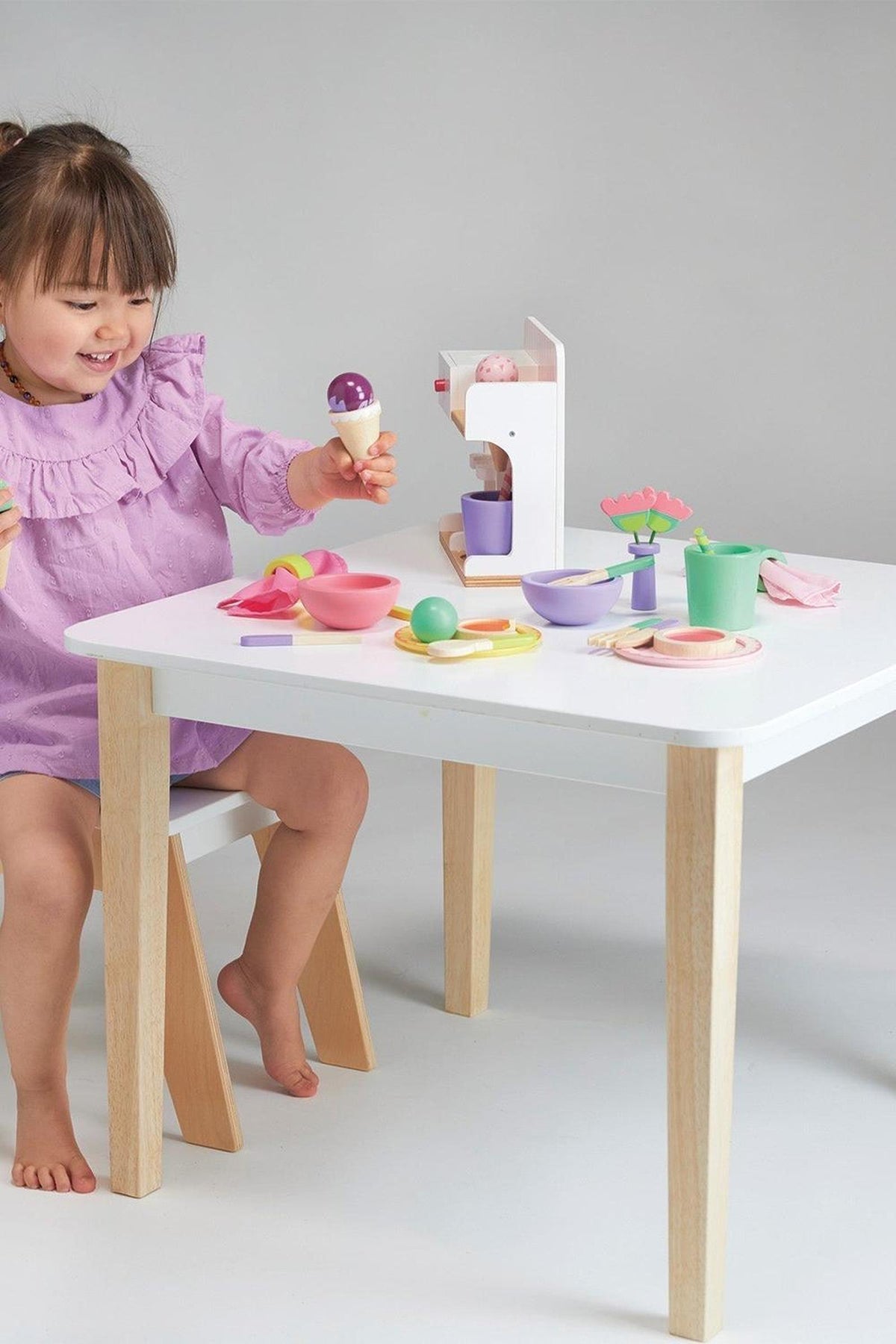 A child in a purple dress plays with a wooden toy lunch set on a white table.