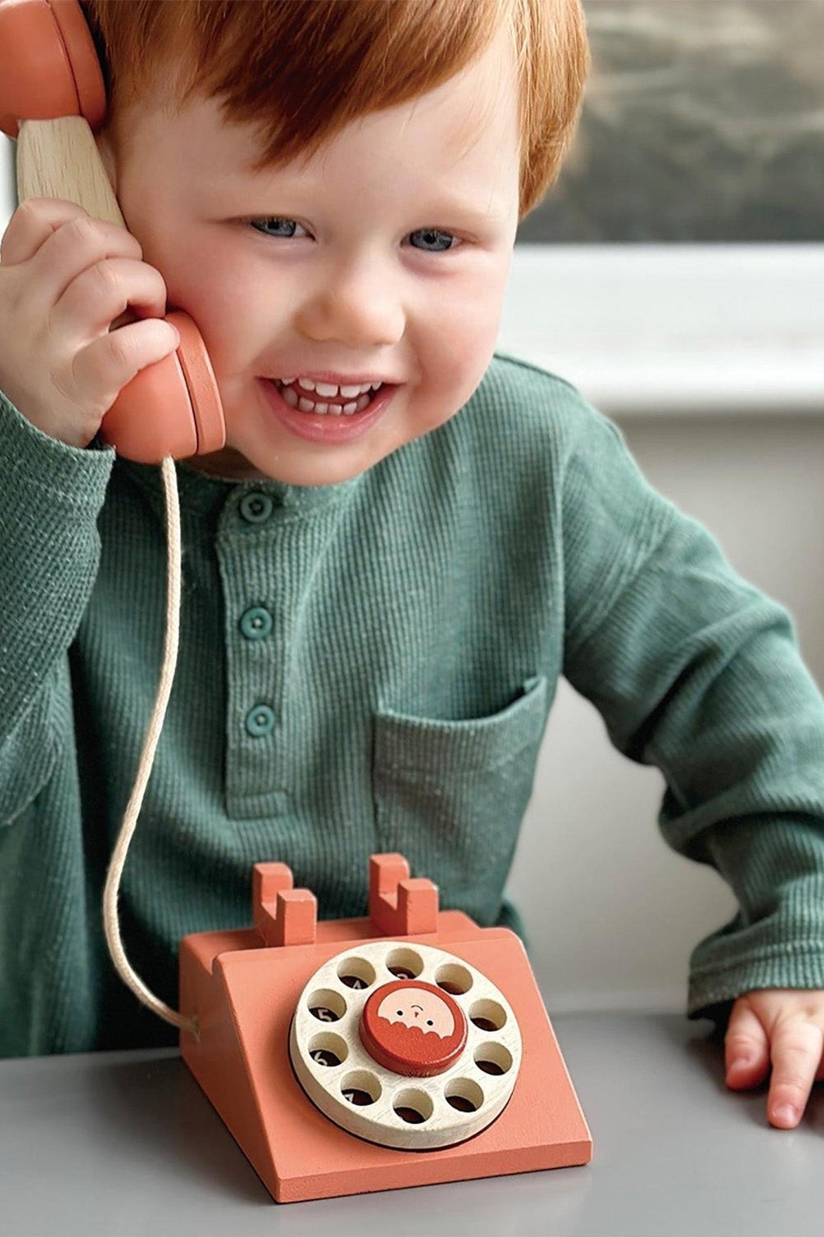 A child with red hair smiles while holding a wooden retro telephone in orange, wearing a green shirt.