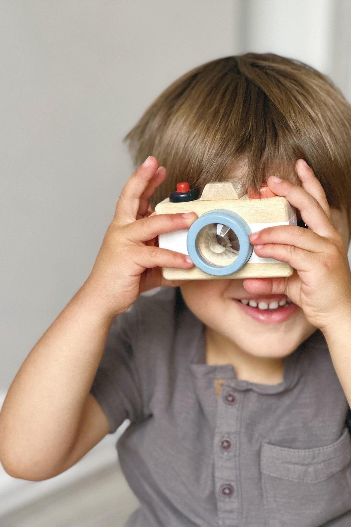 A child holds a wooden toy camera with a blue lens and red button, smiling joyfully indoors.
