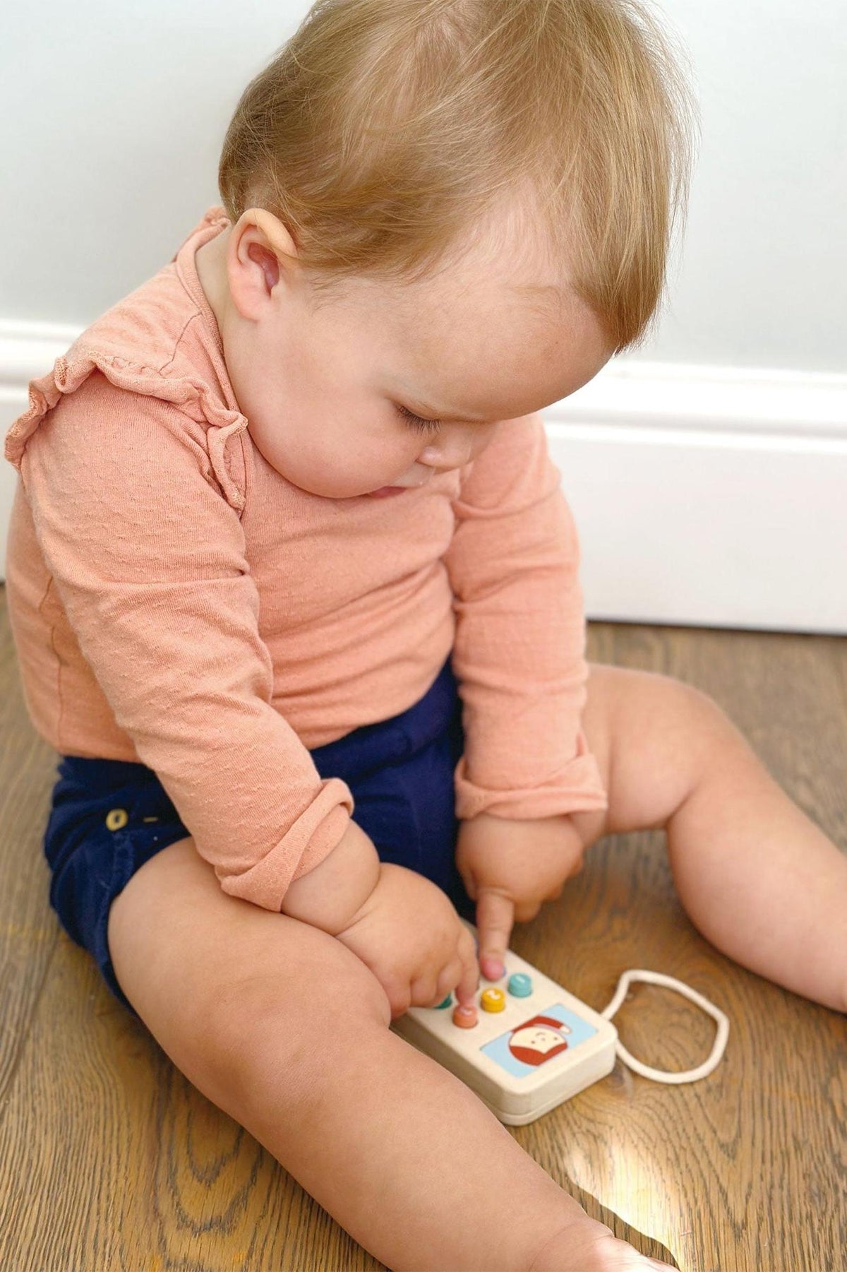 A toddler in a peach top plays with a colorful wooden toy phone on a wooden floor.