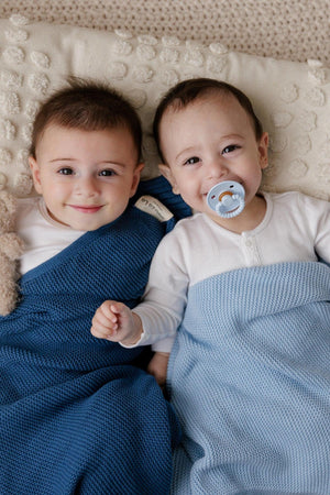 Two smiling babies lie wrapped in blue knit blankets, one with a pacifier, on a textured cream pillow.