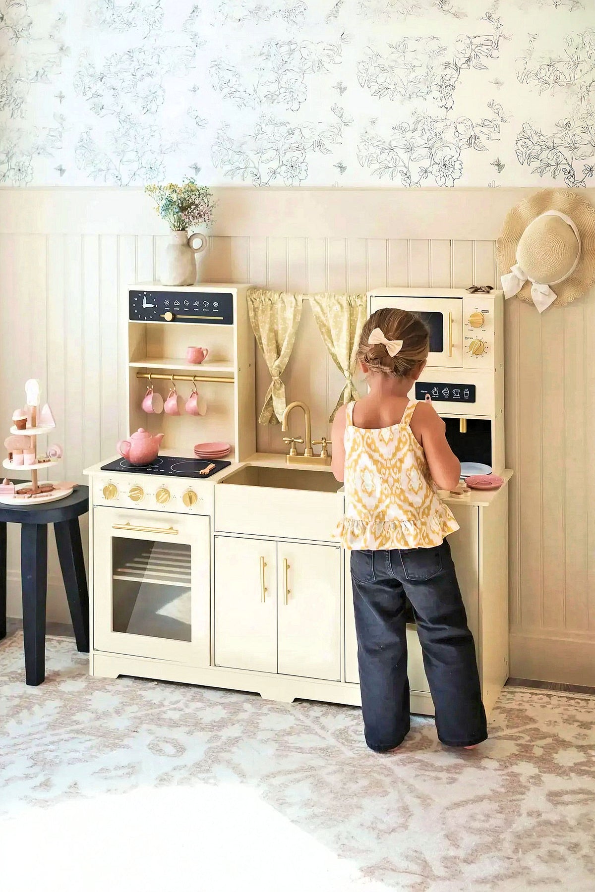 A child stands at a cream-colored wooden play kitchen, featuring realistic appliances like an oven and sink, with decorative curtains and pink dishes, promoting imaginative play.