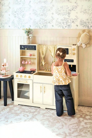 A child stands at a cream-colored wooden play kitchen, featuring realistic appliances like an oven and sink, with decorative curtains and pink dishes, promoting imaginative play.