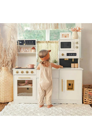 A young child in a beige outfit interacts with the Tiny Land® wooden play kitchen, featuring a cream color, realistic knobs, and various cooking appliances, set against a bright, airy.