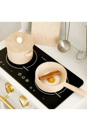 A wooden play kitchen set featuring a cream-colored saucepan and a pot on a black stovetop. The saucepan contains a realistic egg and a piece of bacon, promoting imaginative play.