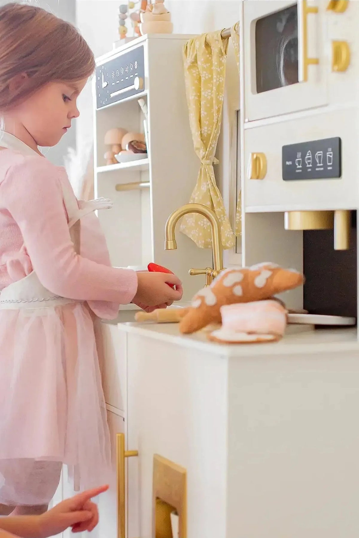 A young girl in a pink dress and apron plays with a wooden play kitchen, featuring realistic details like an oven and microwave. Another child points, engaged in imaginative play.
