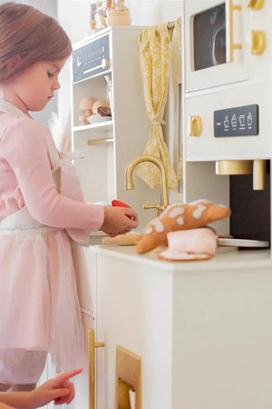 A young girl in a pink dress and apron plays with a wooden play kitchen, featuring realistic details like an oven and microwave. Another child points, engaged in imaginative play.