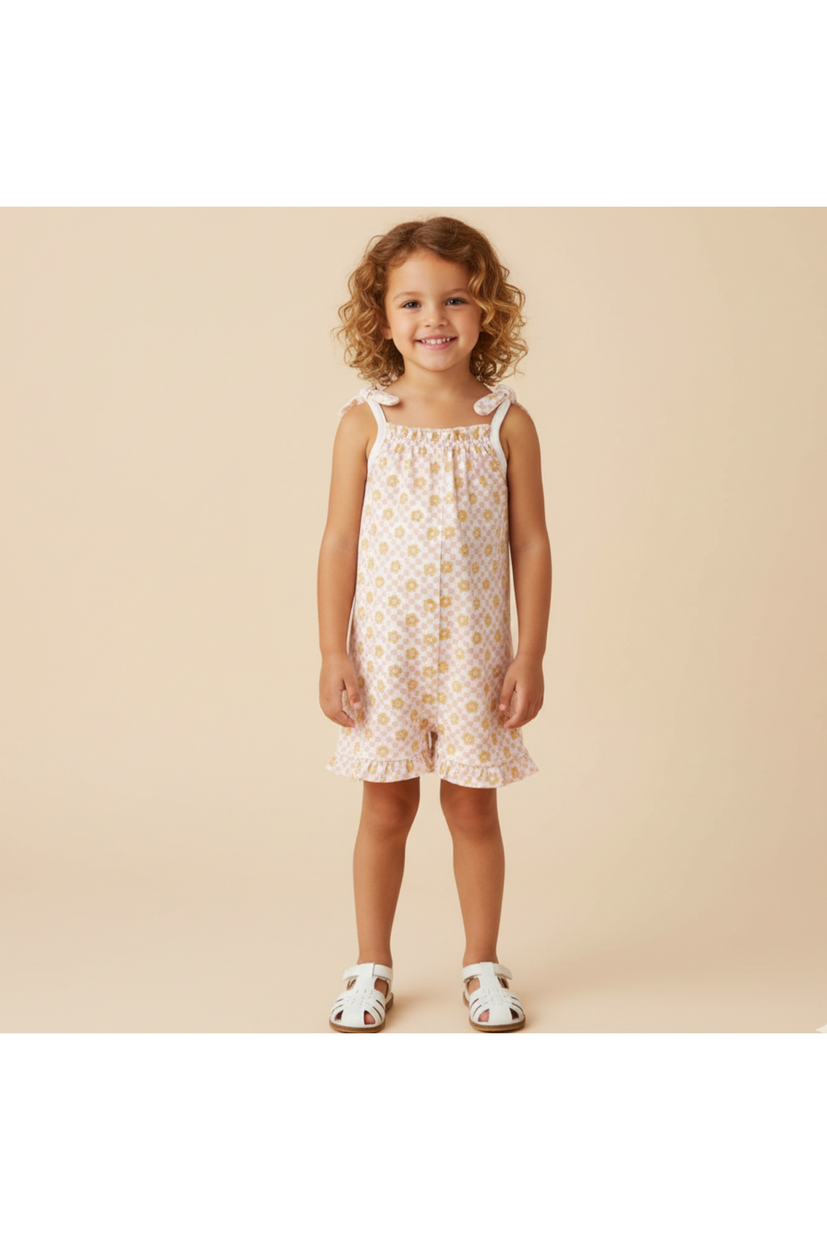 A young girl smiles while wearing a light pink ruffle jumpsuit with a floral pattern. The outfit features adjustable straps and is perfect for sunny days, showcasing a cheerful, playful.