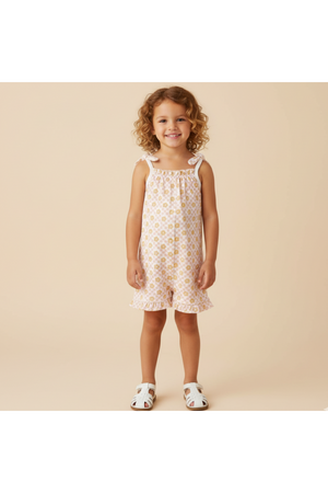 A young girl smiles while wearing a light pink ruffle jumpsuit with a floral pattern. The outfit features adjustable straps and is perfect for sunny days, showcasing a cheerful, playful.