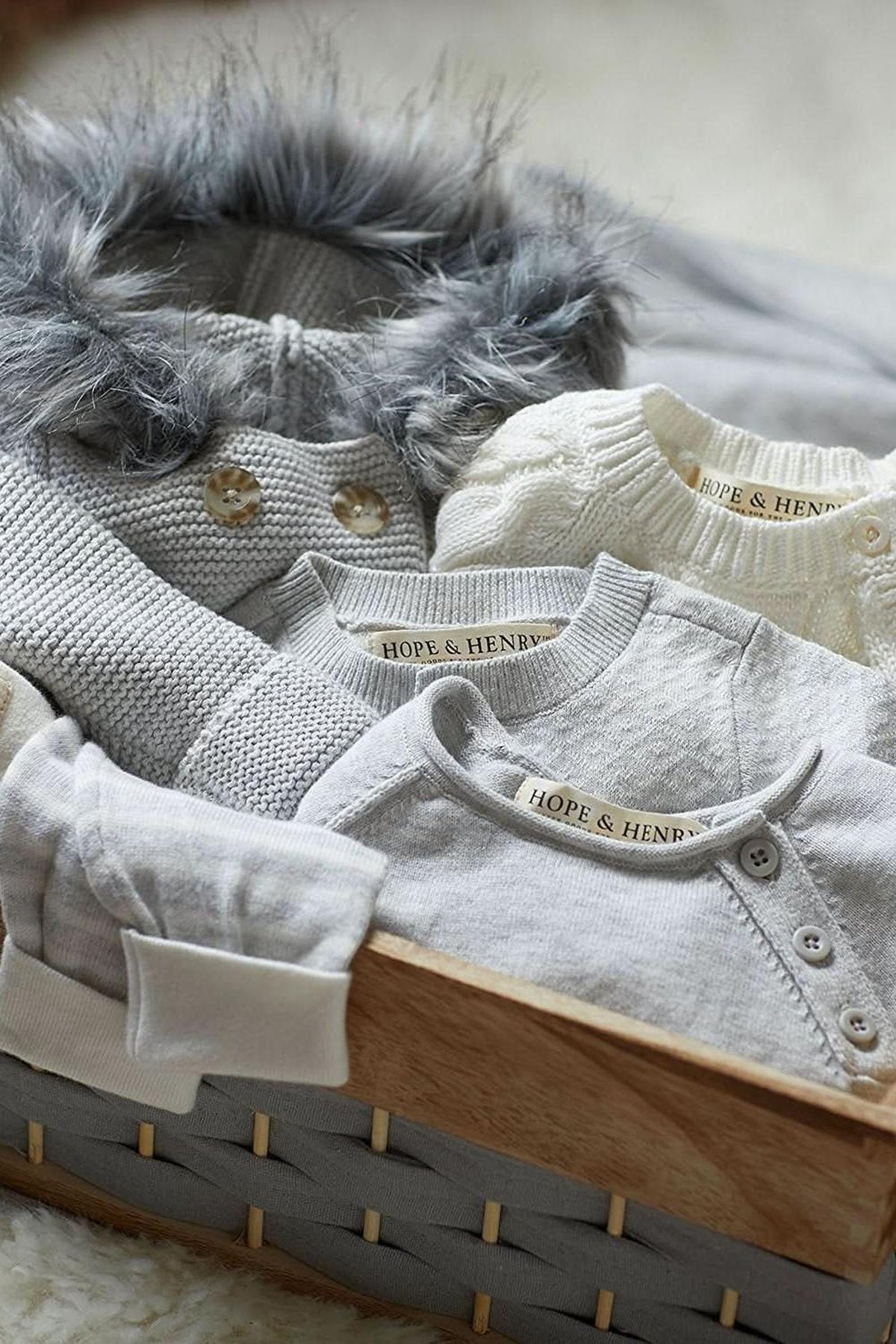 A cozy arrangement of soft, light grey baby garments is displayed in a woven basket. Each piece features the Hope & Henry label, highlighting their organic cotton and sustainable design.