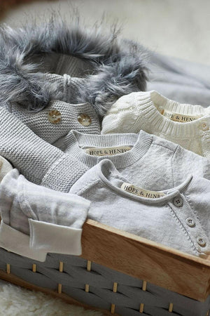 A cozy arrangement of soft, light grey baby garments is displayed in a woven basket. Each piece features the Hope & Henry label, highlighting their organic cotton and sustainable design.