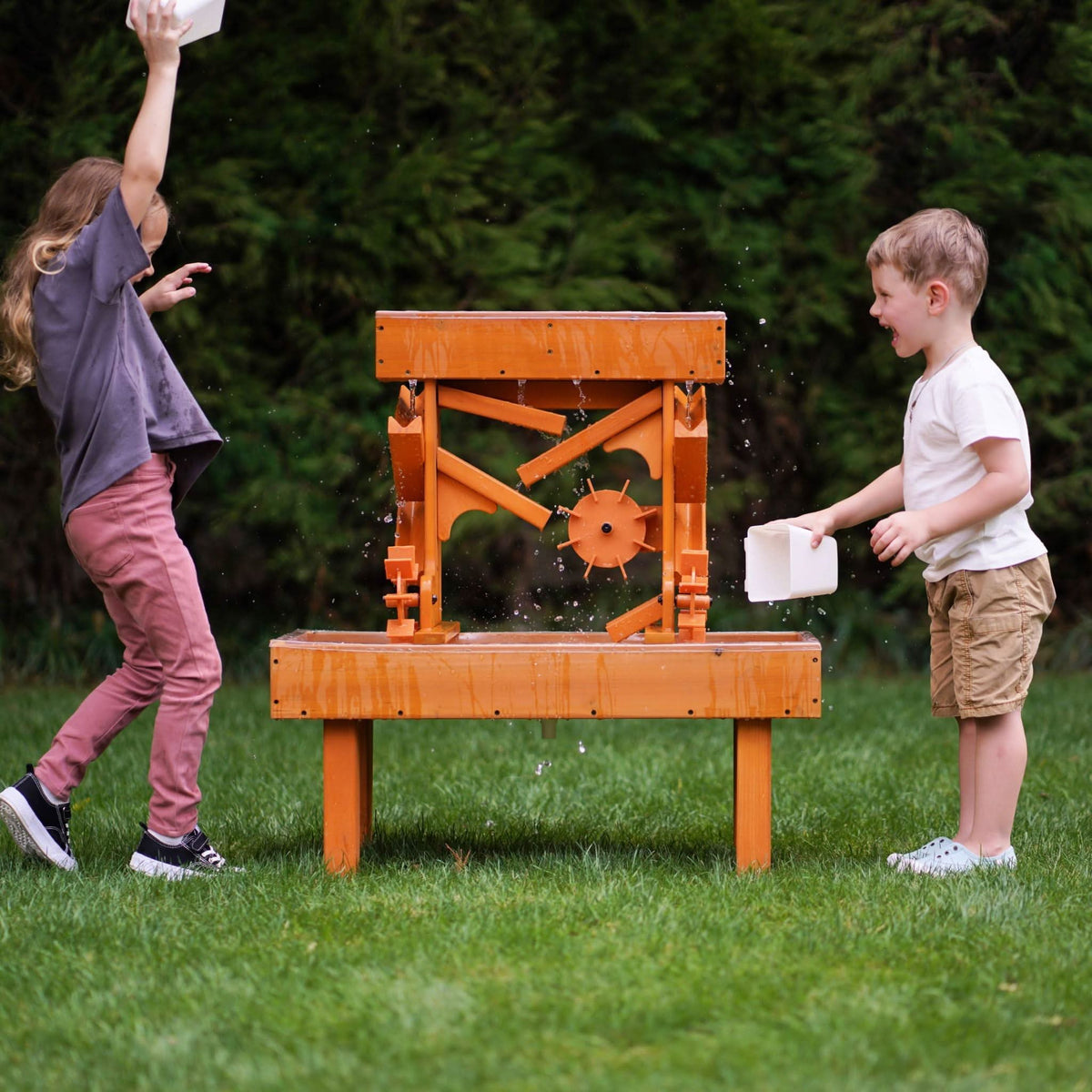 Two children play with an outdoor wooden water table, splashing water joyfully in a grassy setting. The vibrant orange table features channels and a waterwheel for interactive sensory play.