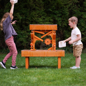 Two children play with an outdoor wooden water table, splashing water joyfully in a grassy setting. The vibrant orange table features channels and a waterwheel for interactive sensory play.