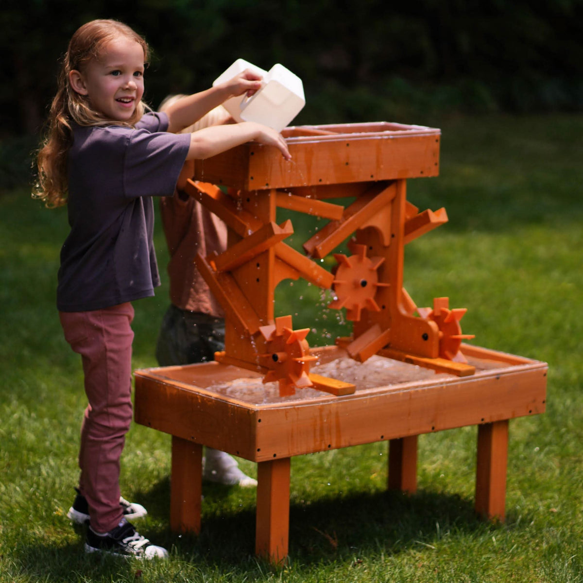 A child with long hair smiles while pouring water from a container into a wooden water table.