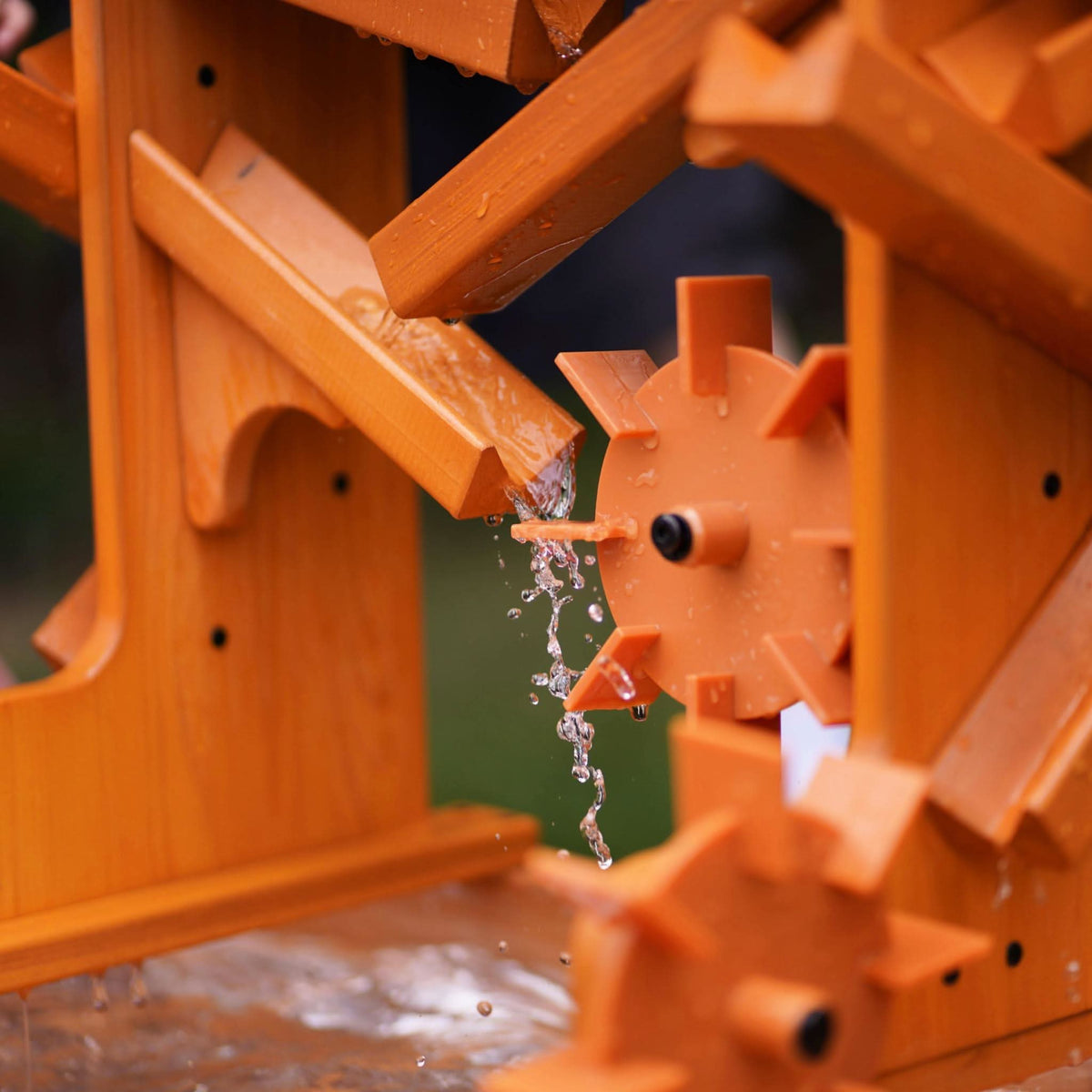 Close-up of a wooden water table featuring vibrant orange gears, with water flowing through channels and splashing. Ideal for sensory play, encouraging exploration and learning through hands-on activities.