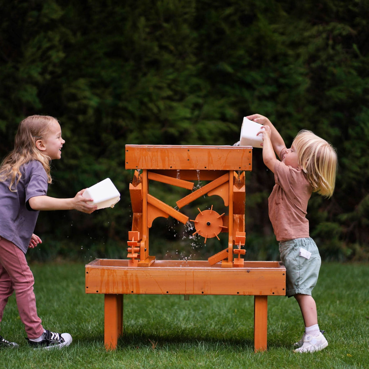 Two children play joyfully with an outdoor wooden water table, featuring vibrant orange construction.