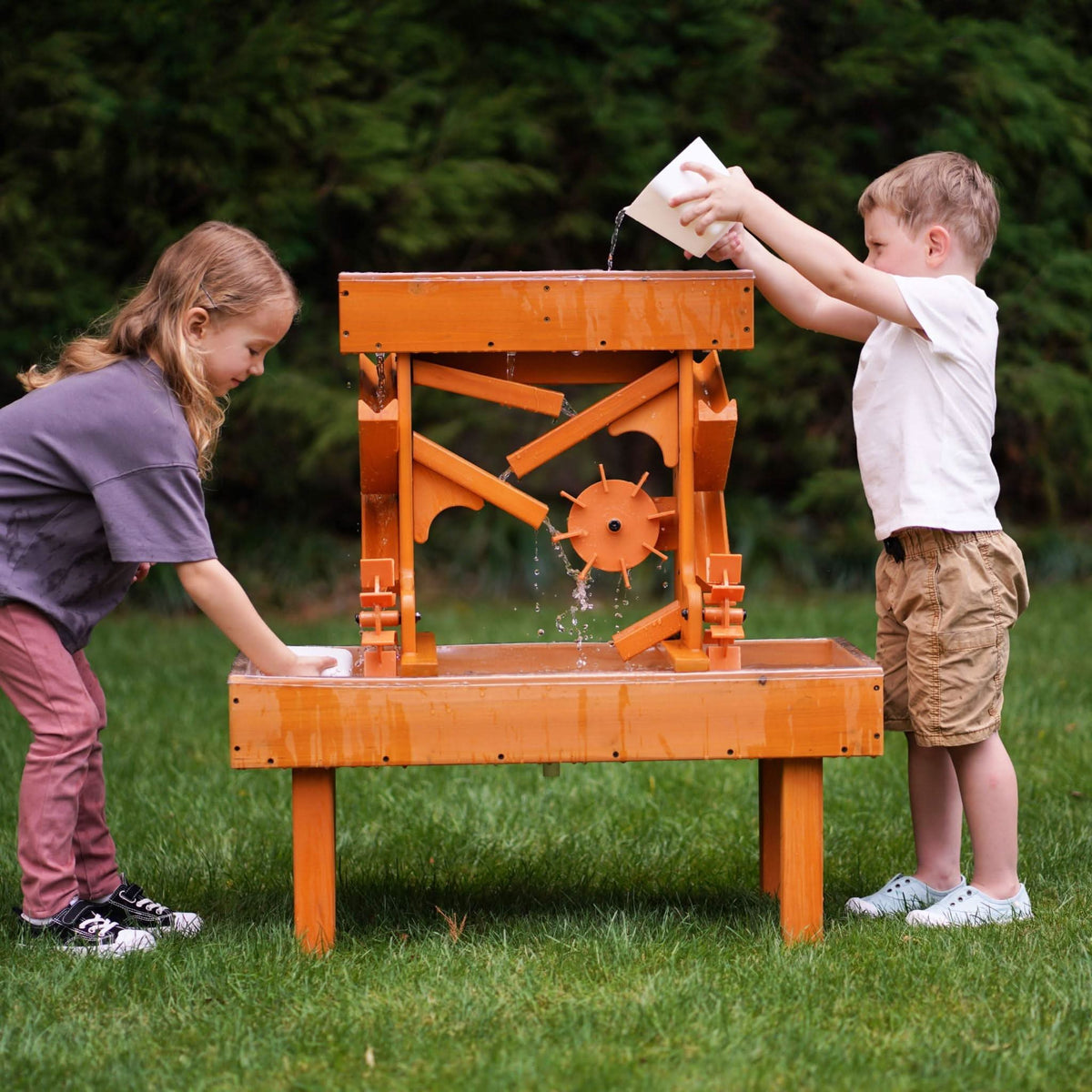 Two children play with an outdoor wooden water table, pouring water and observing its movement through colorful channels and wheels, surrounded by lush green grass.