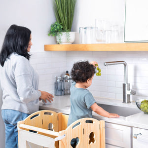 A toddler stands on a wooden Laurel 4-in-1 kitchen tower, reaching for grapes at a modern kitchen sink. An adult is washing dishes nearby, promoting interactive learning and independence.