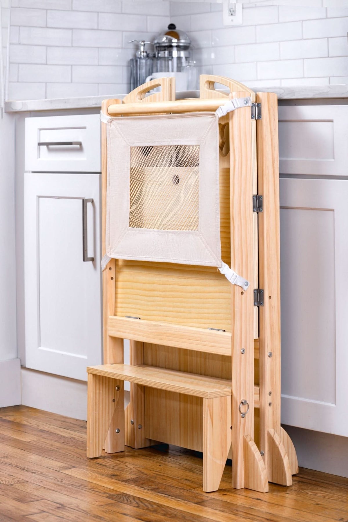 A wooden, foldable kitchen tower features a mesh panel and two-step stool. It is placed against a white cabinet in a bright kitchen, showcasing its versatile design for toddlers.