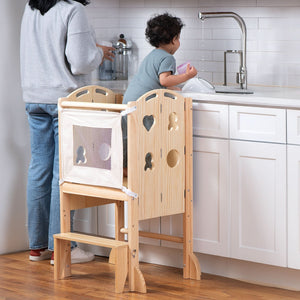 A toddler stands on a natural wood kitchen tower, reaching for a cup at the sink, while an adult prepares food nearby.