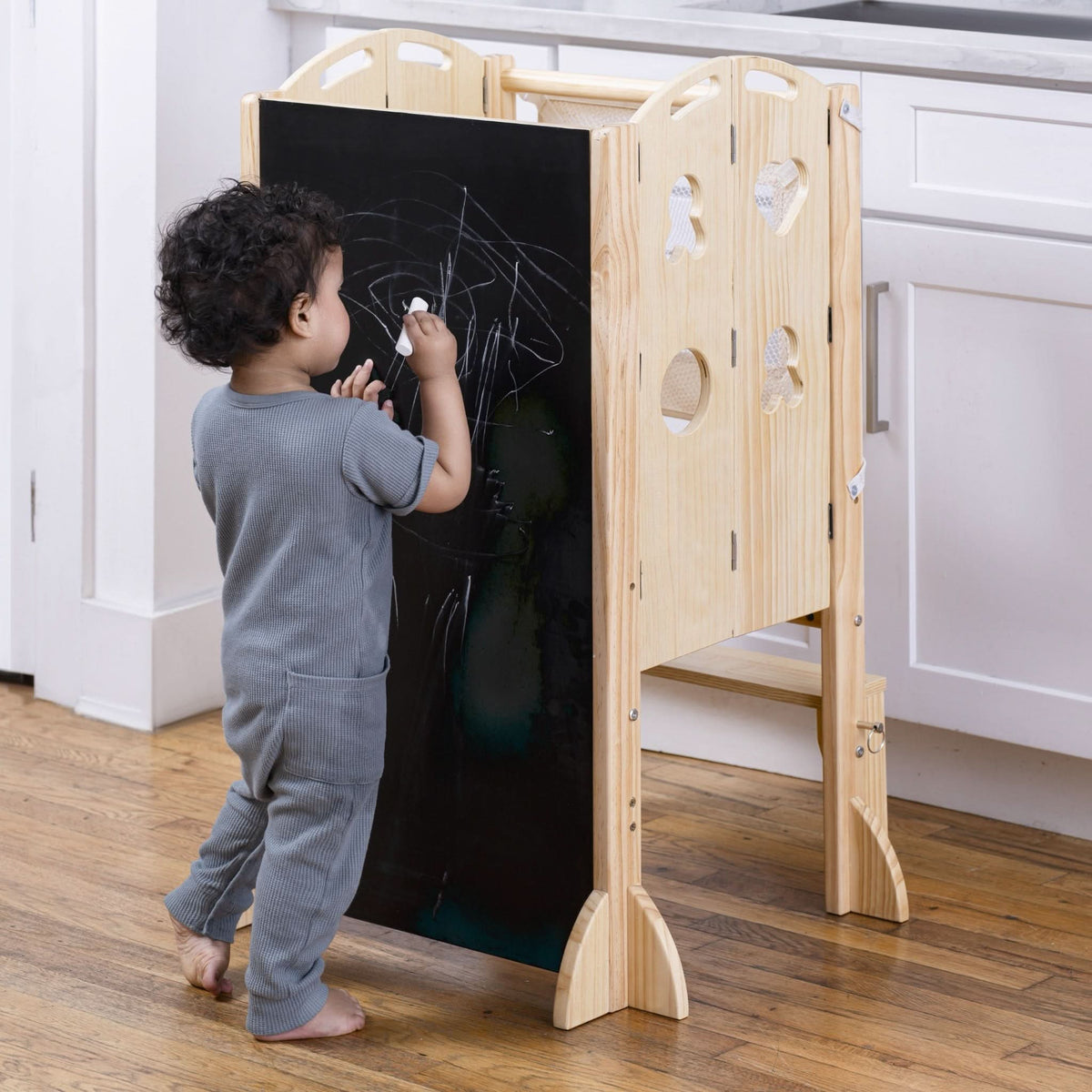 A toddler in gray pajamas uses chalk on a wooden chalkboard attached to a foldable kitchen tower.