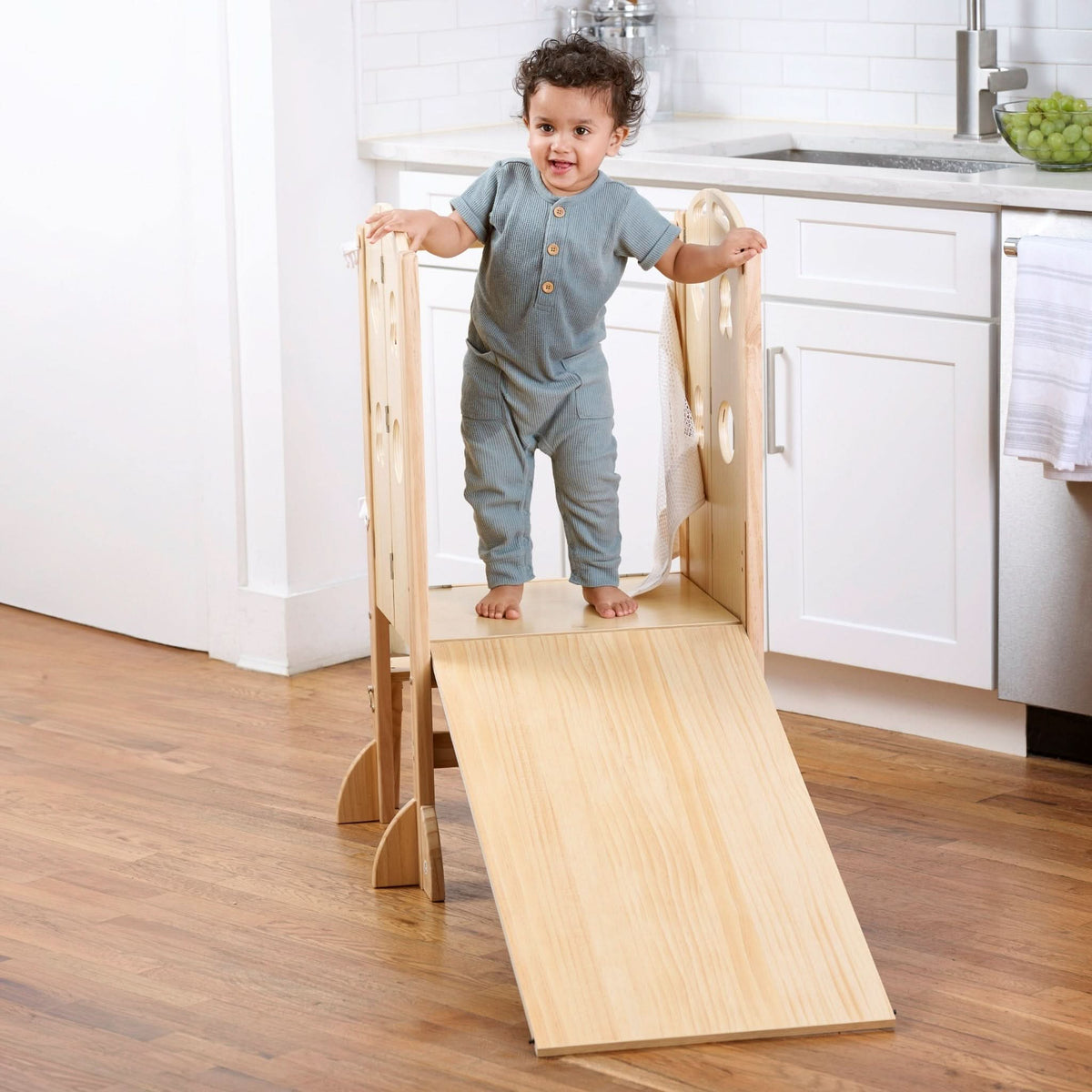 A toddler stands on a wooden foldable kitchen tower with a slide, showcasing a natural wood finish.