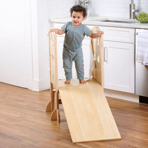 A toddler stands on a wooden foldable kitchen tower with a slide, showcasing a natural wood finish.