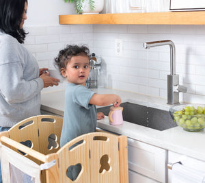 A toddler stands in a wooden kitchen tower, pouring water from a pink pitcher into a sink, while a woman washes dishes nearby.