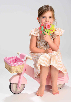 A child sits on a pink wooden scooter with a wicker basket, holding colorful flower toys, wearing a checkered dress.