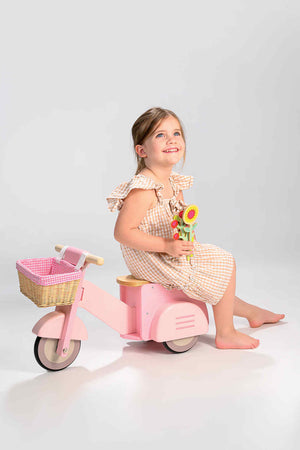 A smiling child sits on a pink wooden scooter with a wicker basket, holding flowers, against a light background.