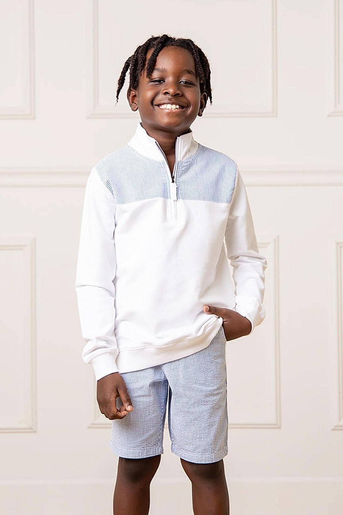 A smiling boy wears a white zip-up sweatshirt paired with blue and white seersucker shorts, showcasing a modern fit and playful style. The background features a light, neutral wall.