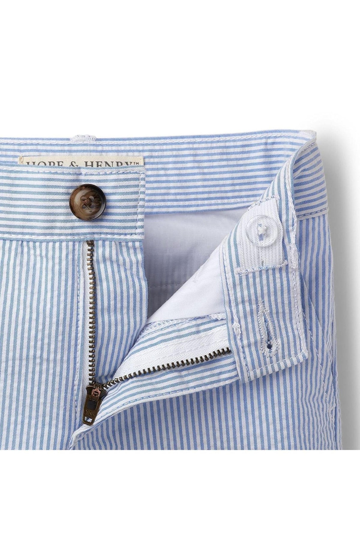Close-up view of blue seersucker shorts featuring an adjustable waistband, a faux horn button, and a zipper. Made from lightweight organic cotton, these shorts are designed for comfort and style.