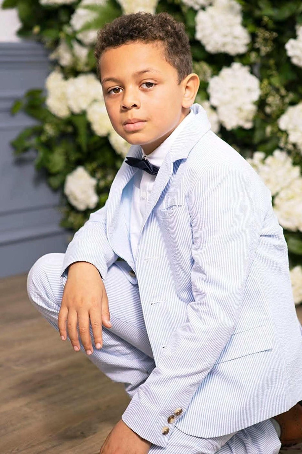 A young boy poses in a light blue seersucker suit jacket, featuring a classic notch collar and button front, against a backdrop of white flowers.