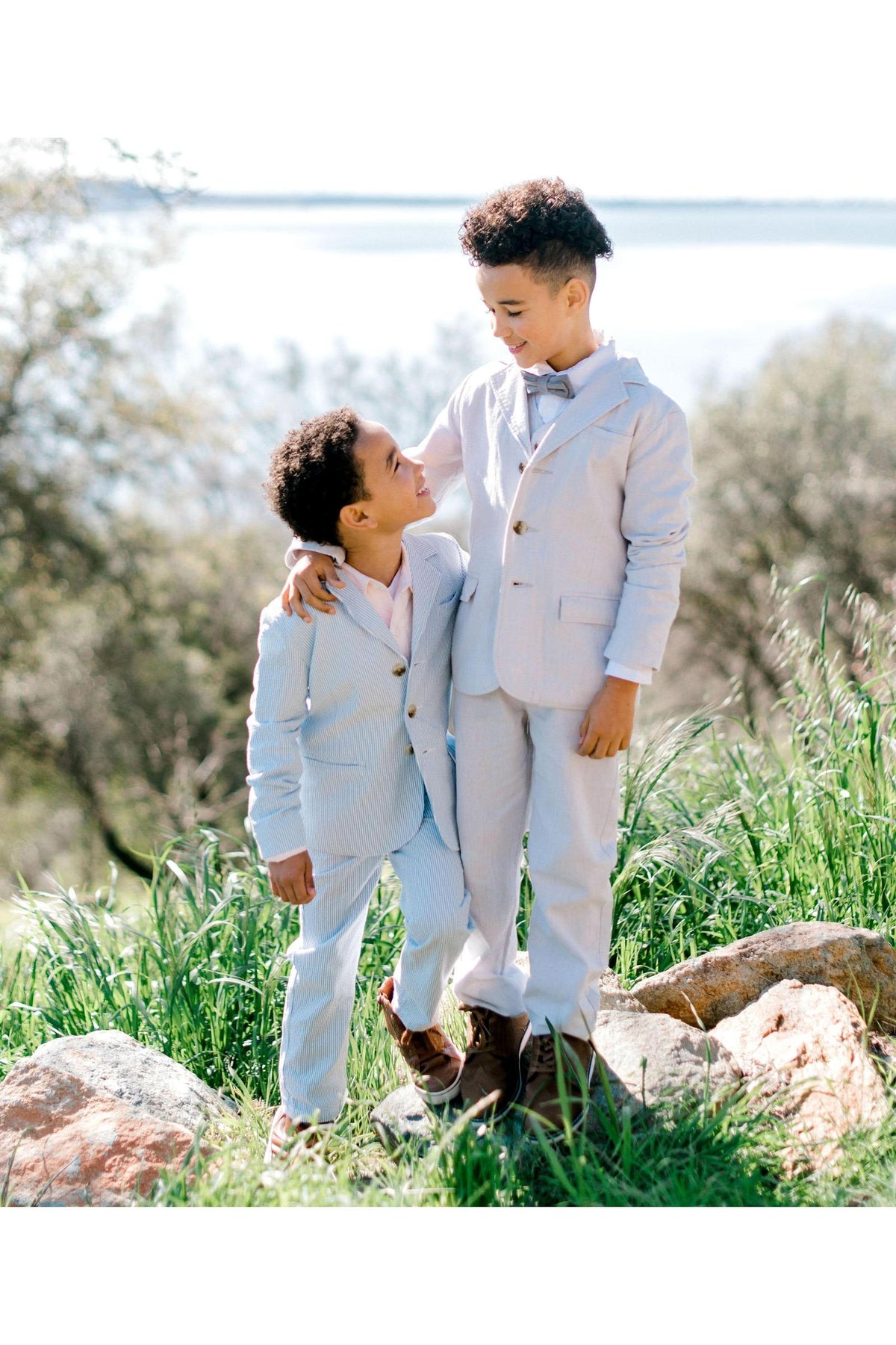 Two boys stand outdoors, one wearing a light blue seersucker suit and the other in a matching jacket and pants, both styled for a special occasion with a scenic backdrop.