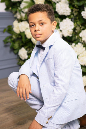 A young boy in a light blue seersucker suit crouches against a backdrop of white flowers. The suit is styled with a bow tie, highlighting a polished, classic look.
