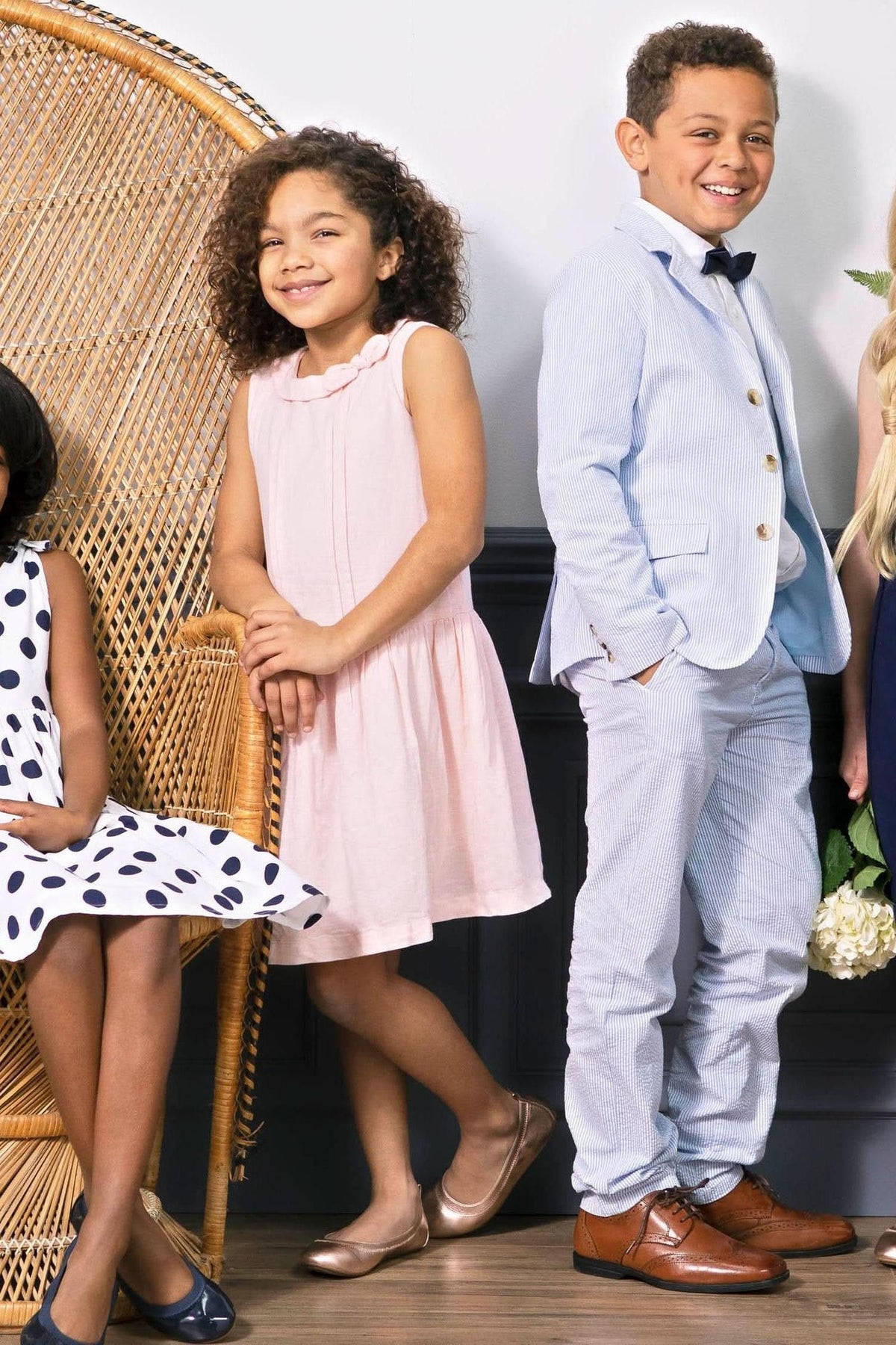 A boy in a light blue seersucker suit pant and bow tie stands confidently beside a girl in a pink dress, both smiling against a stylish, woven backdrop.