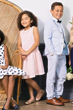 A boy in a light blue seersucker suit pant and bow tie stands confidently beside a girl in a pink dress, both smiling against a stylish, woven backdrop.