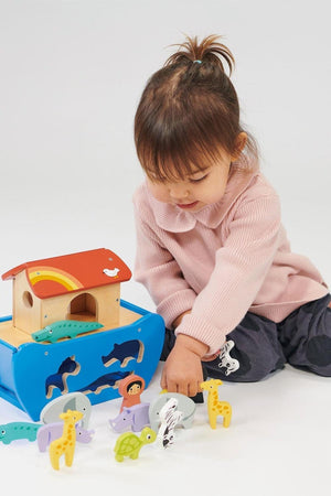A child in a pink sweater plays with a colorful wooden animal ark toy set on the floor.