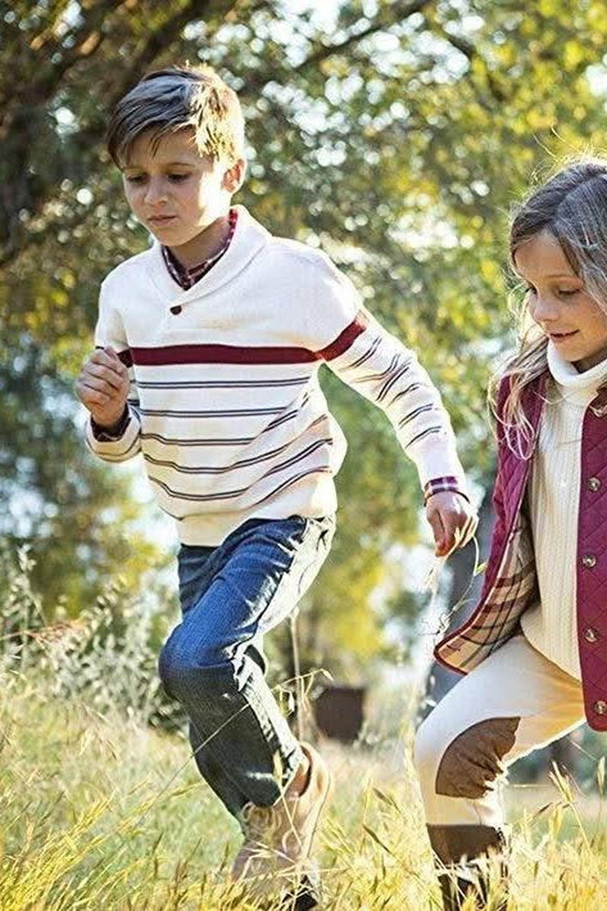 Two children run through a sunlit field, wearing a striped ivory sweater and a quilted jacket, enjoying the outdoors.