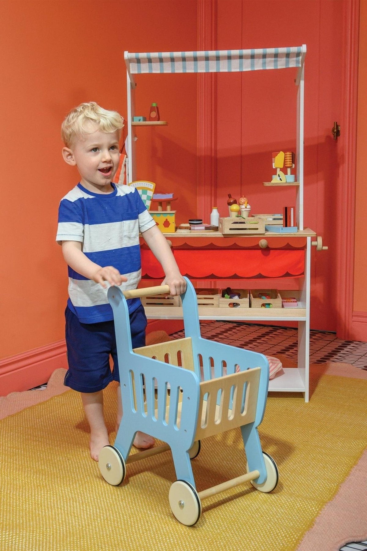 A child pushes a blue wooden shopping cart in front of a colorful toy market stand with play items.