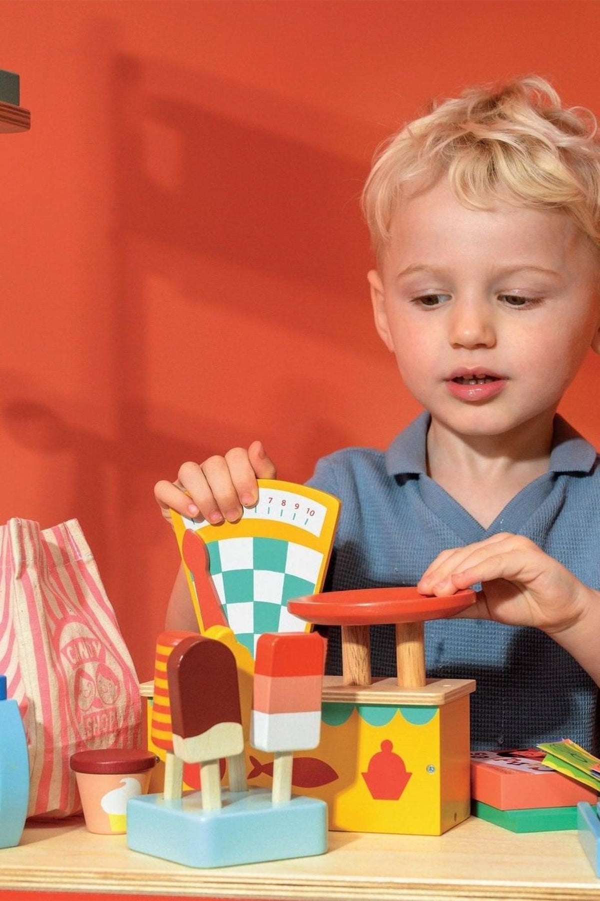 A child plays with colorful wooden toy market items, including pretend ice creams and a scale, against an orange background.