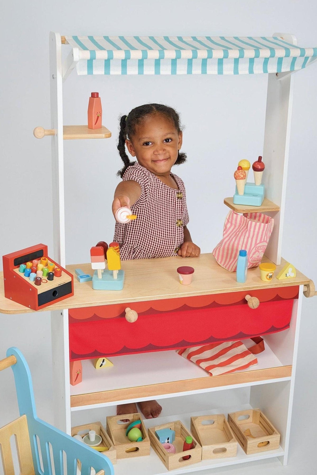 A child plays with the Mentari Wooden Toys Shop Keeper Stand, featuring colorful wooden items and a striped canopy.