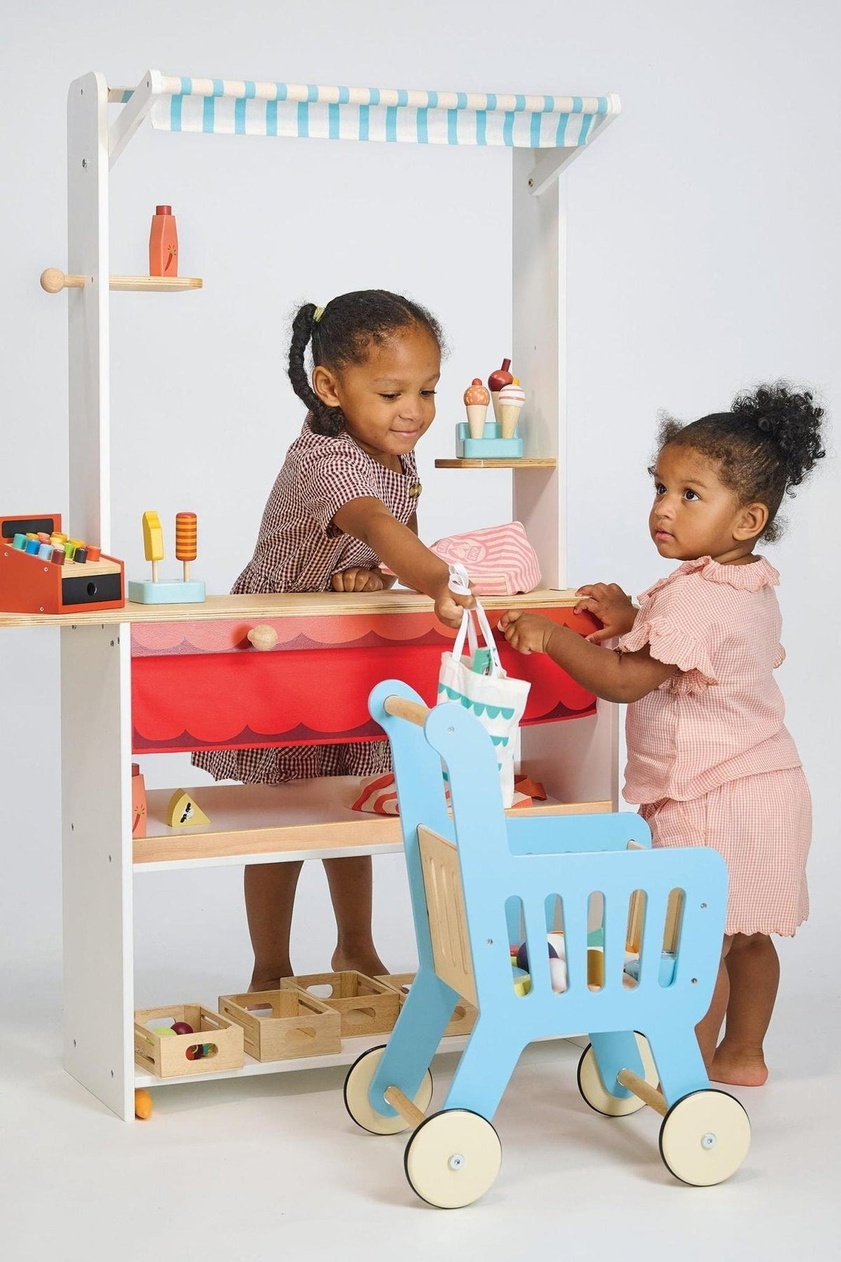Two children play with a wooden shopkeeper stand, featuring a blue-striped canopy and a toy shopping cart.