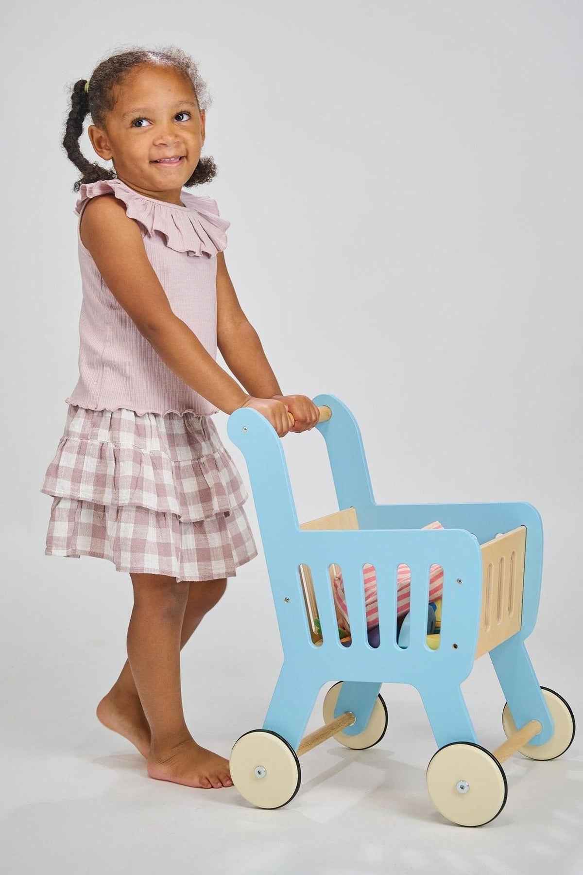 A child in a pink outfit pushes a blue wooden toy shopping cart on a white background.