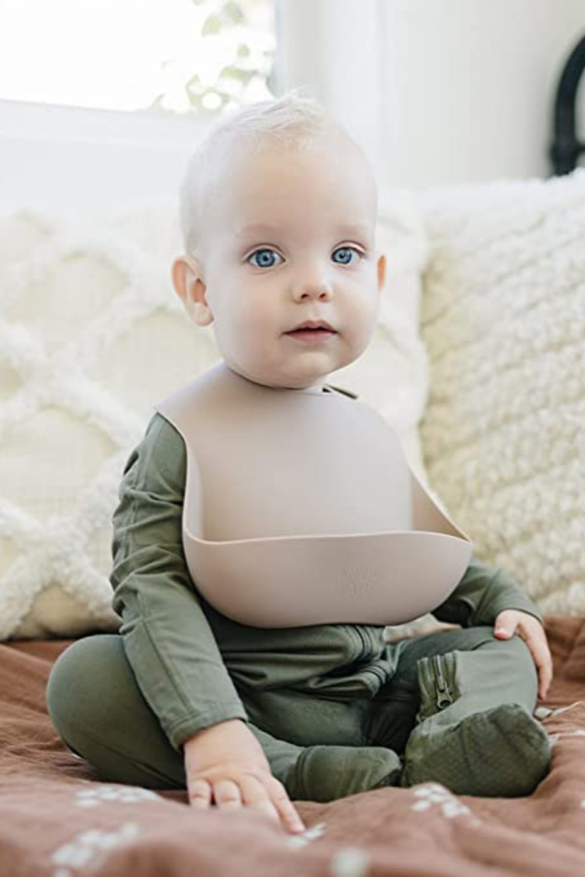 A baby wearing a beige silicone bib and green outfit sits on a brown blanket in a cozy room.