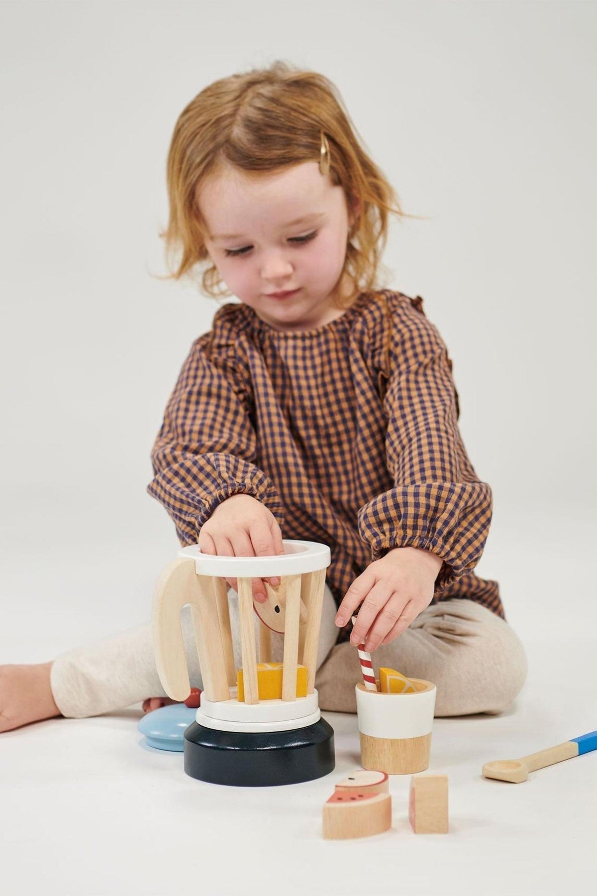 A child in a brown checkered dress plays with a wooden smoothie maker toy set on a white background.
