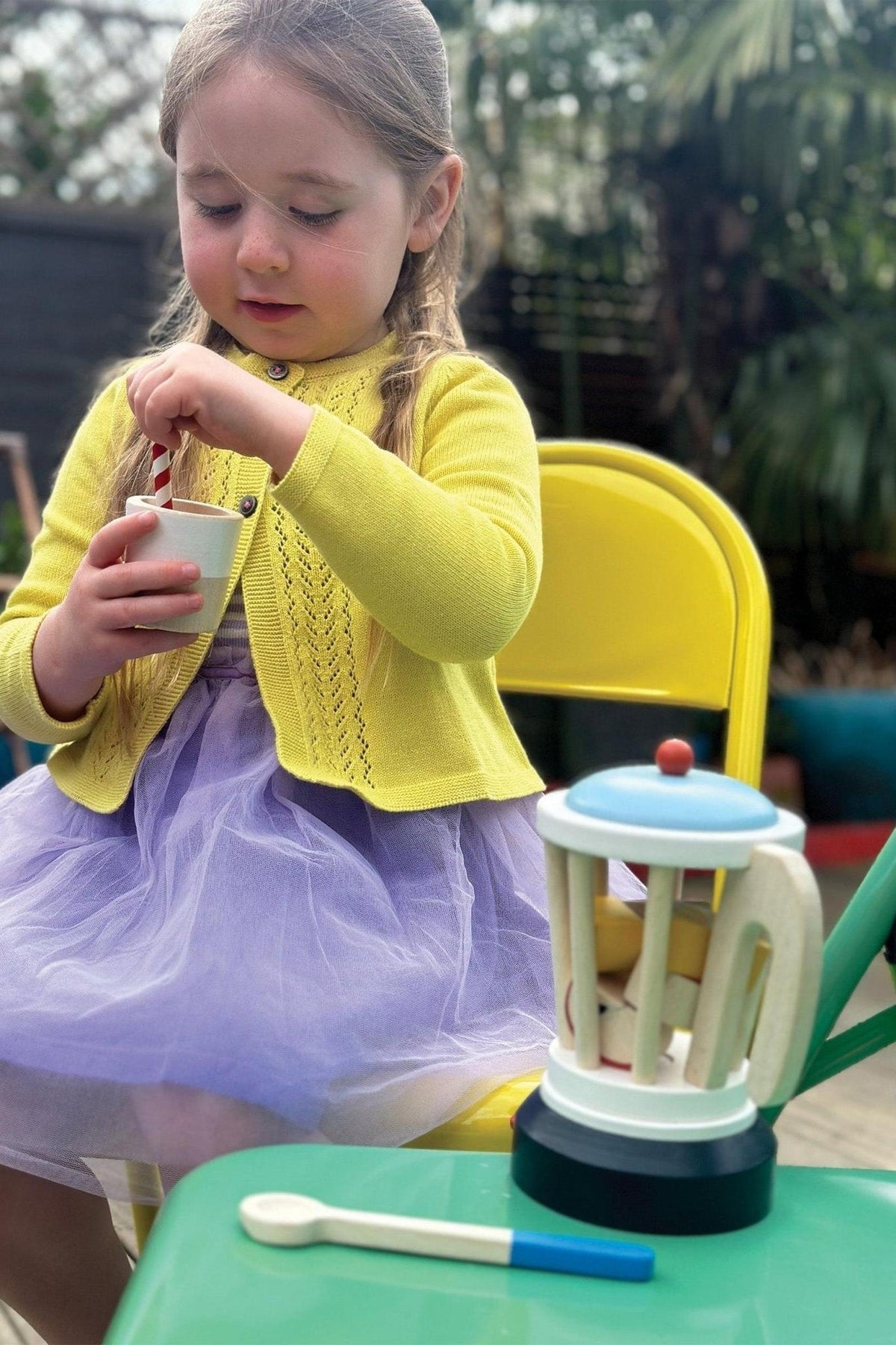 A child in a yellow sweater and purple skirt plays with a wooden smoothie set on a green table.